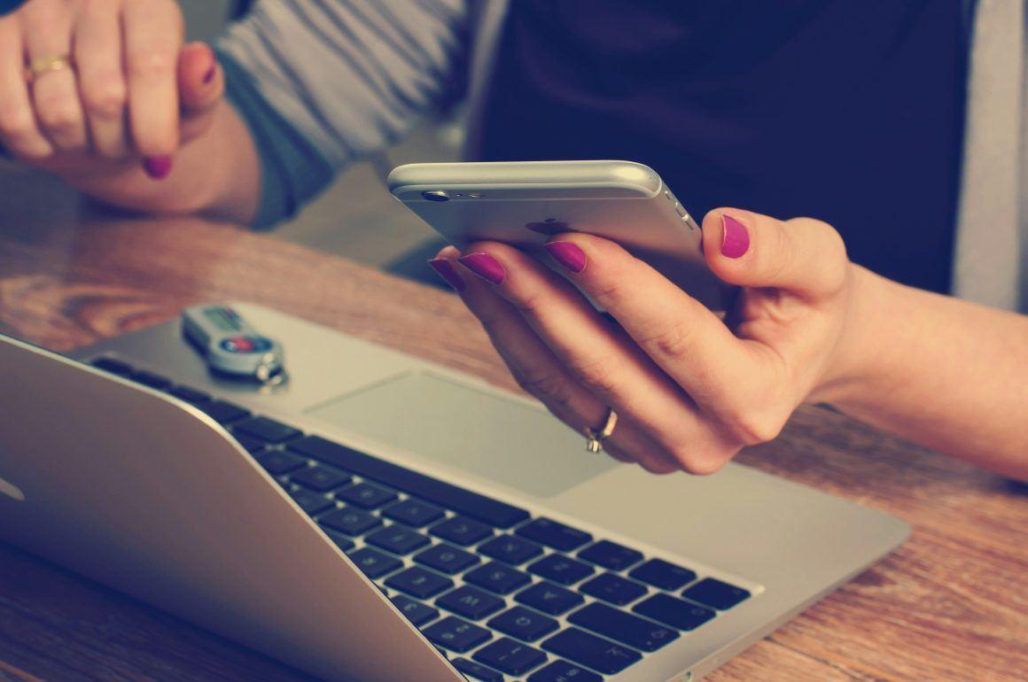 An image of a woman with two forms of technology on the go looking at phone and using laptop