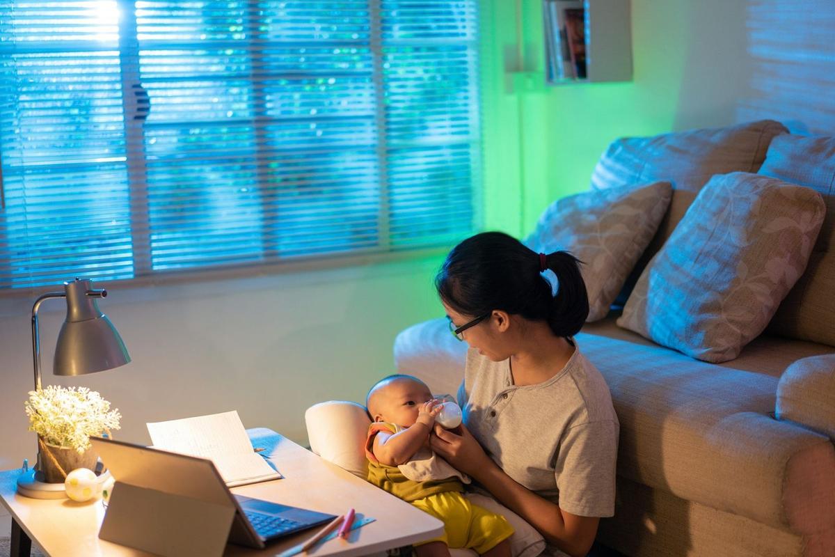 An image of woman sat in her lounge on the floor with her baby in her arms whilst she is working at the coffee table with her laptop and papers around her