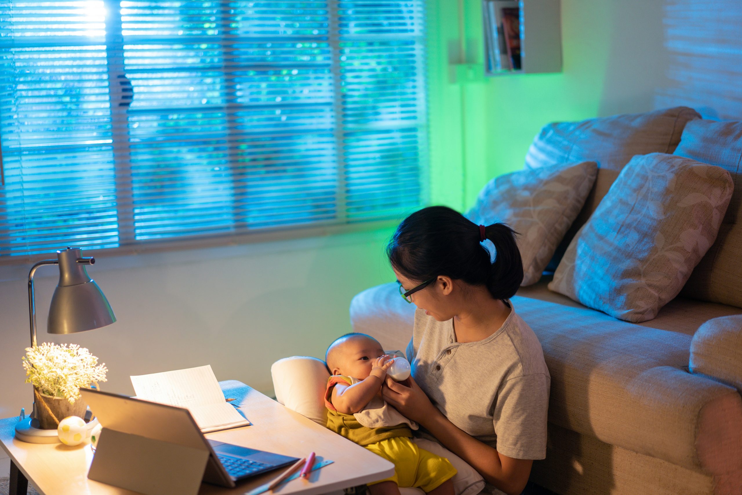 An image of woman sat in her lounge on the floor with her baby in her arms whilst she is working at the coffee table with her laptop and papers around her